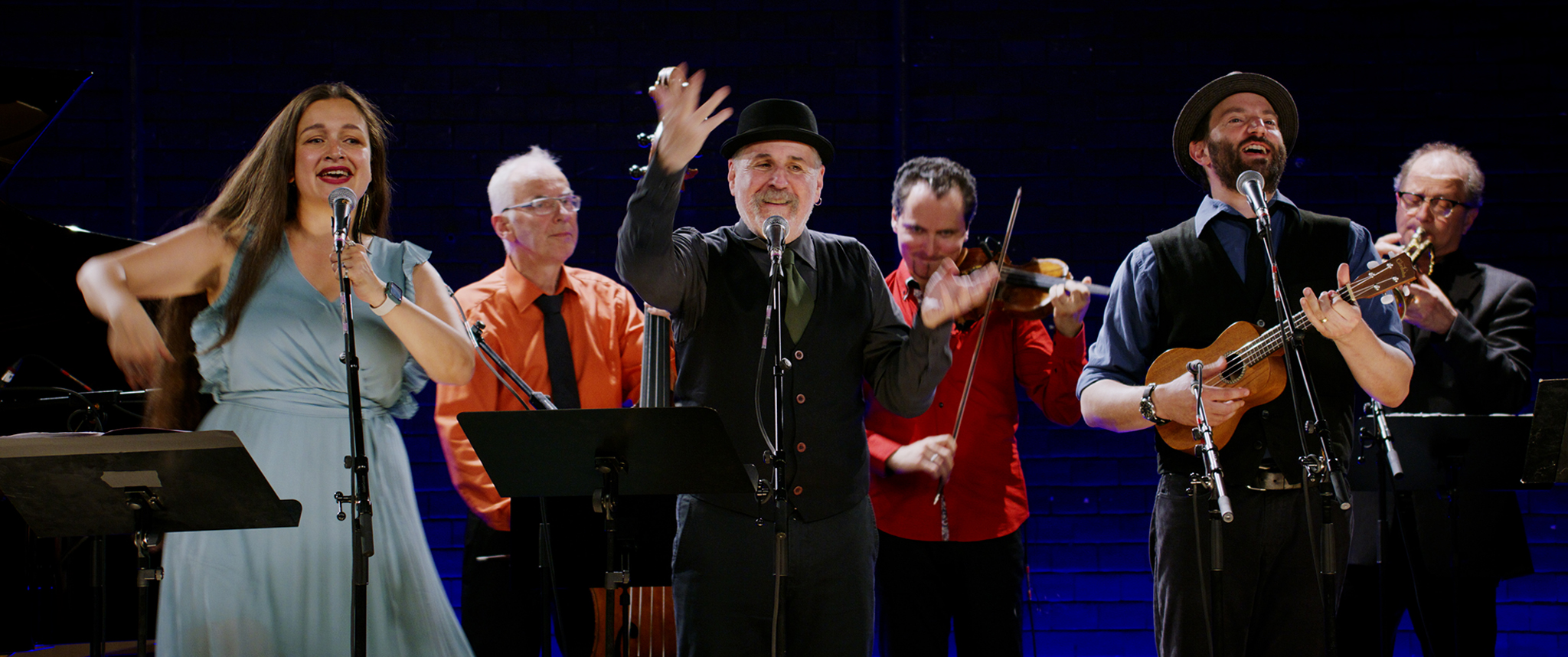 Eine Musikgruppe tritt auf, bestehend aus mehreren Sänger*innen und Musikern, die verschiedene Instrumente spielen. Die Atmosphäre ist lebhaft und locker.