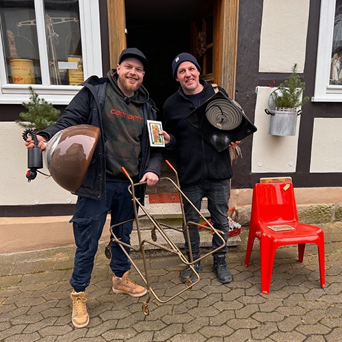 Zwei Männer stehen mit Vintagemöbeln vor einem alten Fachwerkhaus