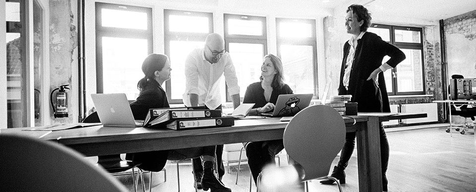 Vier Personen in einem modernen Büro sitzen an einem Tisch, arbeiten zusammen und diskutieren mit Laptops und Ordnern.