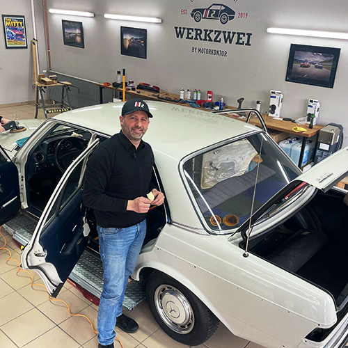 Mann steht in Werkstatt mit weißem Oldtimer